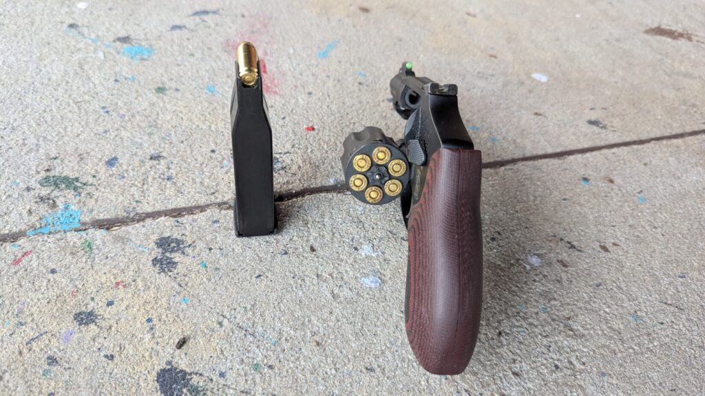 A rear view of a Smith & Wesson revolver with an open cylinder next to a slim semi-auto magazine, highlighting the width of the cylinder versus a single-stack magazine.