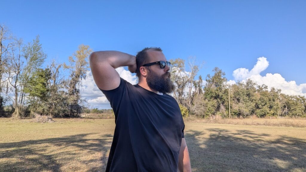 A man in sunglasses and a beard reaching behind his head, demonstrating a "grooming gesture" or a reach for a concealed weapon at the neckline.