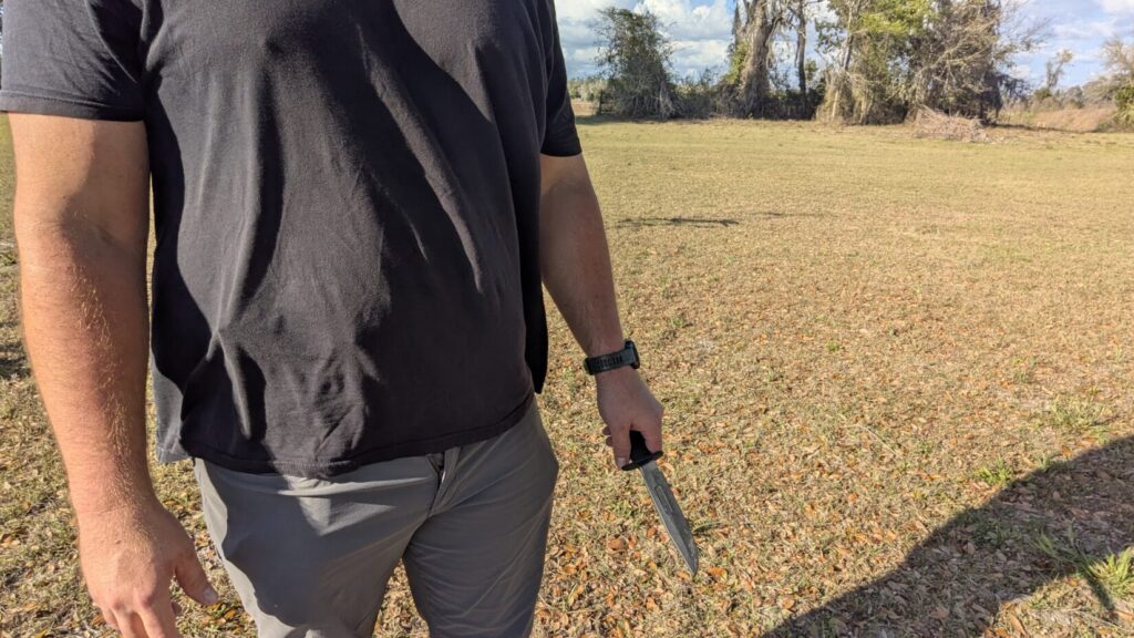 A close-up of a man’s hand holding a fixed-blade knife pointed downward, kept close to the thigh to minimize visibility.