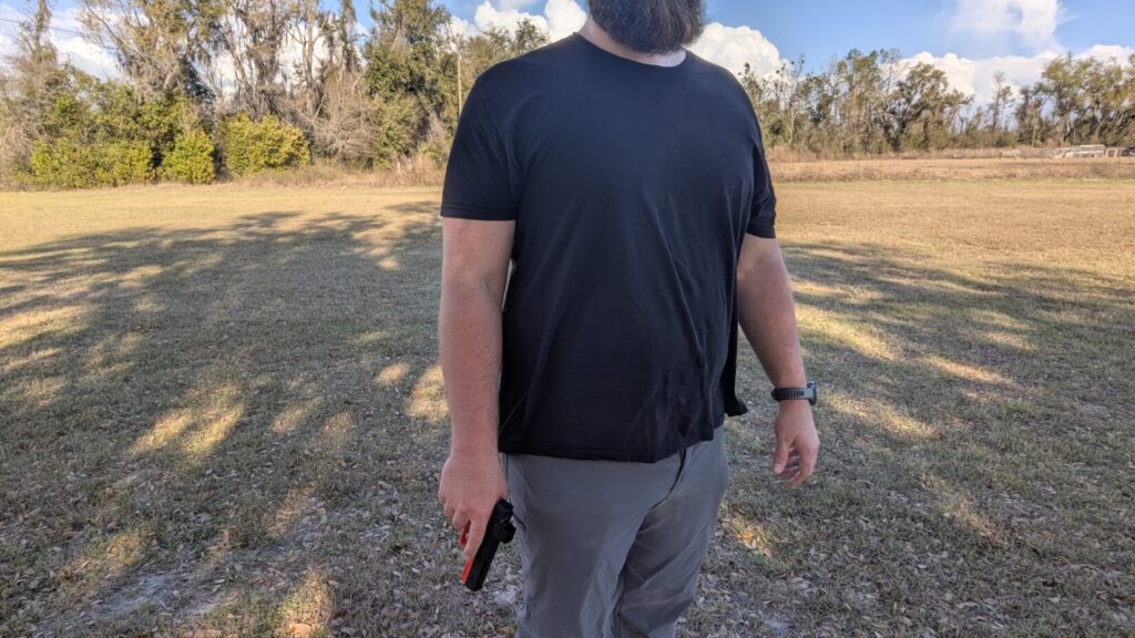 A man standing with a handgun held low and pressed against the side of his leg to hide it from a frontal view.