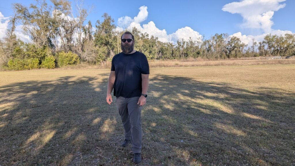 A man standing in an open field with a wide, bladed stance and hands held away from the body, indicating a "ready" or combative posture.
