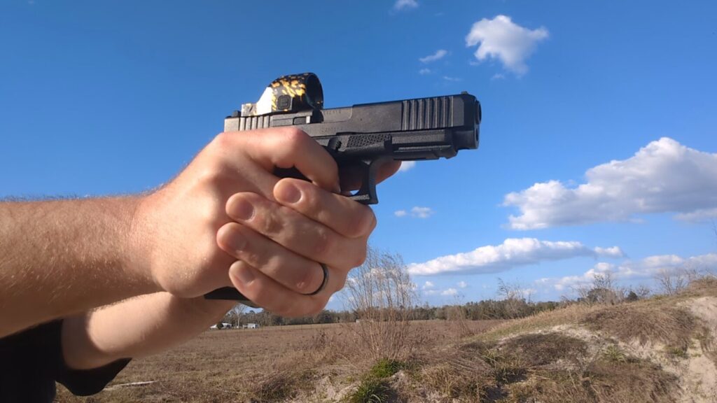 Close-up of a person's hands holding a black semi-automatic pistol with a yellow-patterned red dot sight against a blue sky.