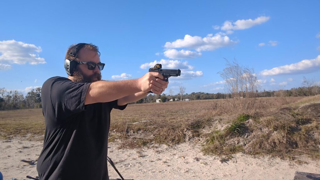 Profile view of a man with a beard and sunglasses aiming a black pistol with a patterned red dot sight in an open field.