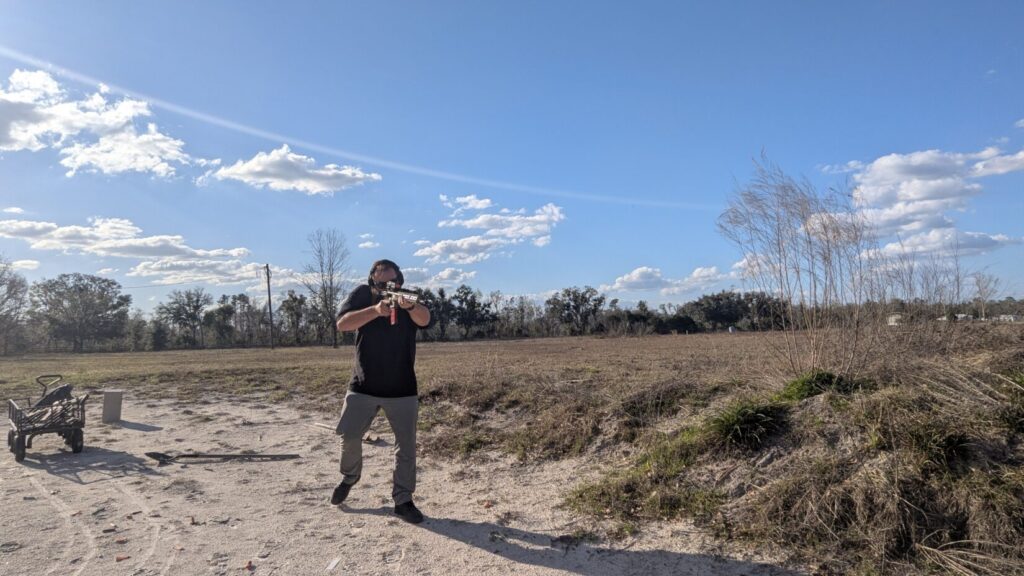  A wide shot of a person standing in a dirt field aiming a tan rifle toward the camera.