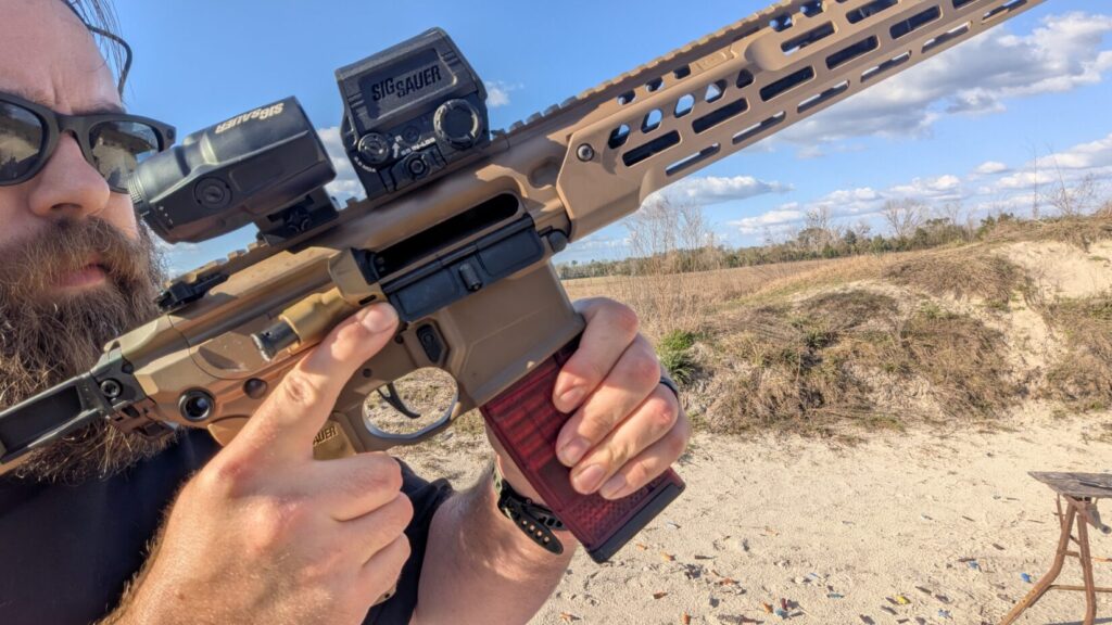 Detailed close-up of a tan Sig Sauer rifle, showing the receiver, red dot optic, and a red-tinted translucent magazine.
