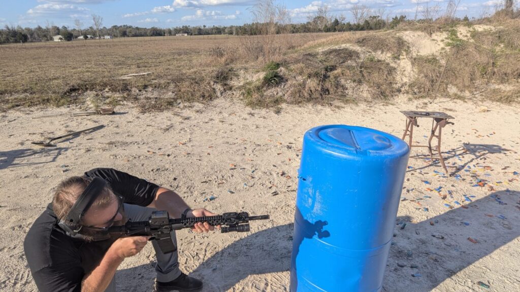 A person kneeling and aiming a black rifle at a blue plastic barrel in an open field littered with spent shells.