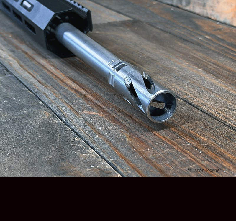 The end of a stainless steel barrel on a rifle, laying on a wood table.