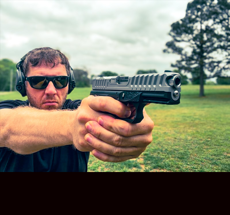 A man in a black shirt and sunglasses, shooting a silver and black handgun on the range. 