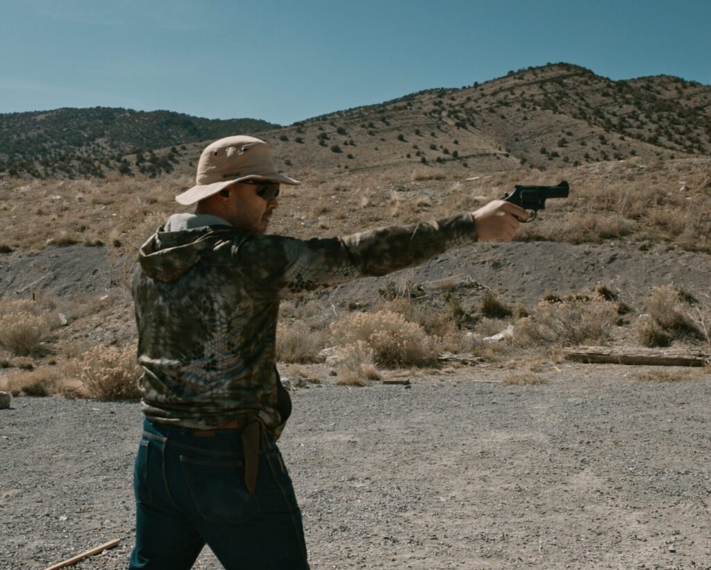 A man in camouflage and wide-brim hat takes aim with a revolver with one hand in a desert setting.