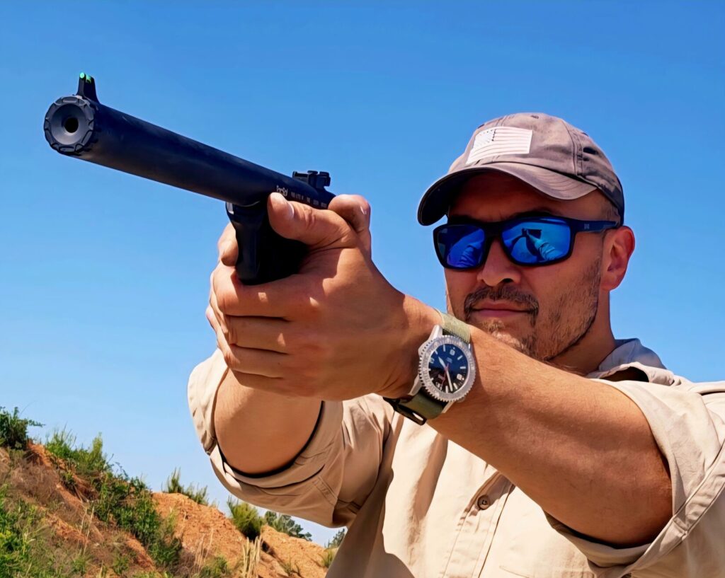 A man in a baseball hat and tan shirt takes aim with a pistol