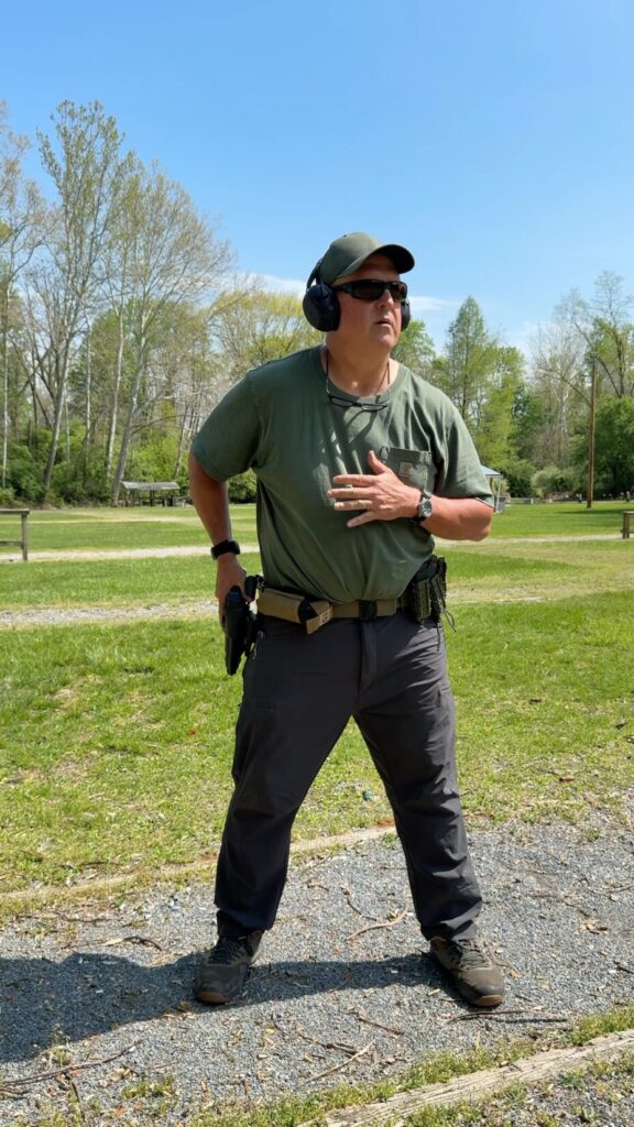 shooter in a green hat, green t shirt and grey pants with his hand on a holstered pistol