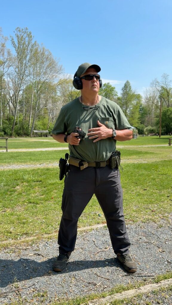 shooter in a green hat, green shirt, grey pants holding a gun in his right hand, pointing at the target after just clearing the holster.