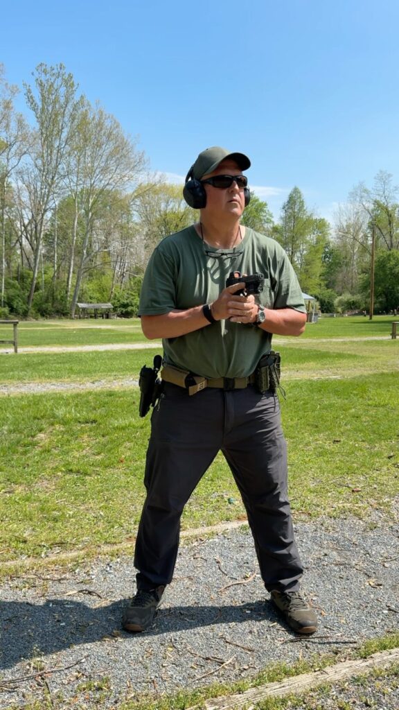 man in a green hat, green t shirt, grey pants, holding a pistol in his right hand mating his left hand with the gun hand during the draw stroke