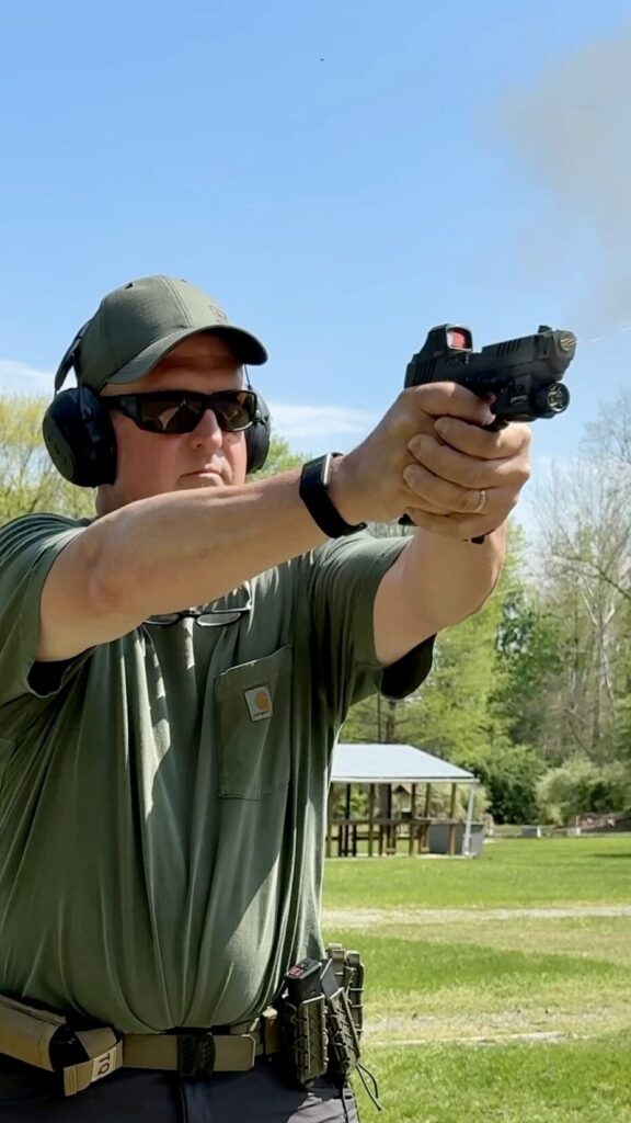 a man in a green hat, green t shirt holding a handgun in a two hand grip. a shot has just been fired and smoke is exiting the barrel.