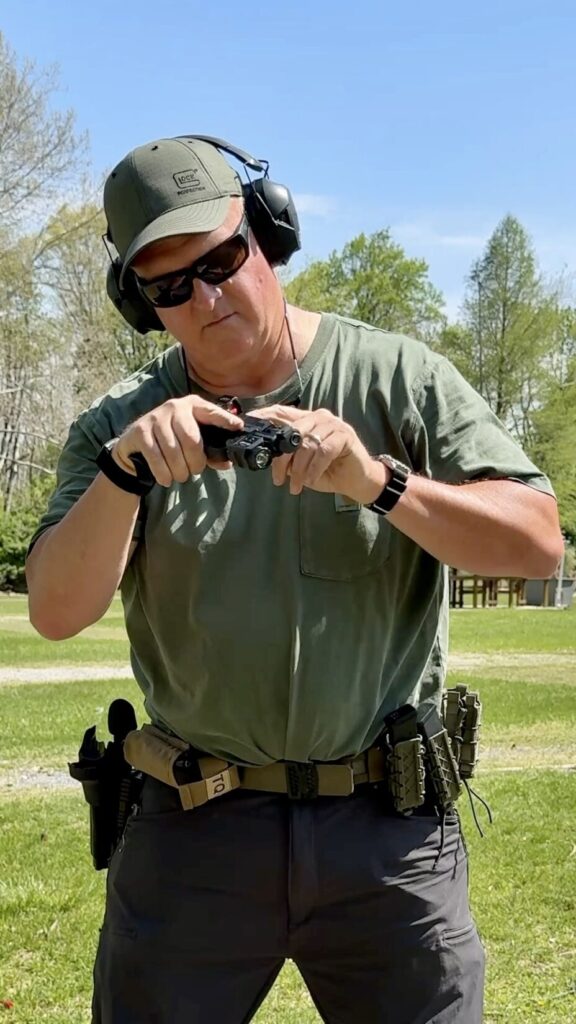 a man in a green hat wearing a green t shirt and grey pants is visually and physically inspecting a handgun with the slide locked to the rear between strings of fire.