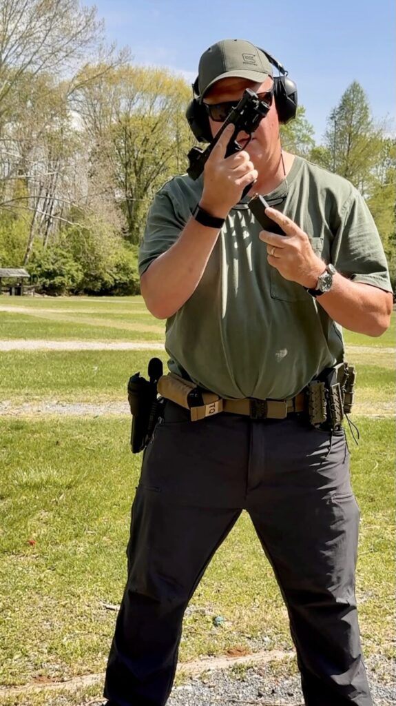 a man in a green hat, green shirt and grey pants holding a pistol in his right hand with the slide locked to the rear. His left hand is bringing a magazine into contact with the grip of the pistol