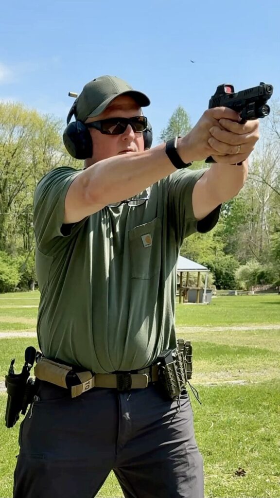 a man in a green hat, green t shirt and grey pants, holding a black pistol in a two handed grip. 2 pieces of brass are in the air