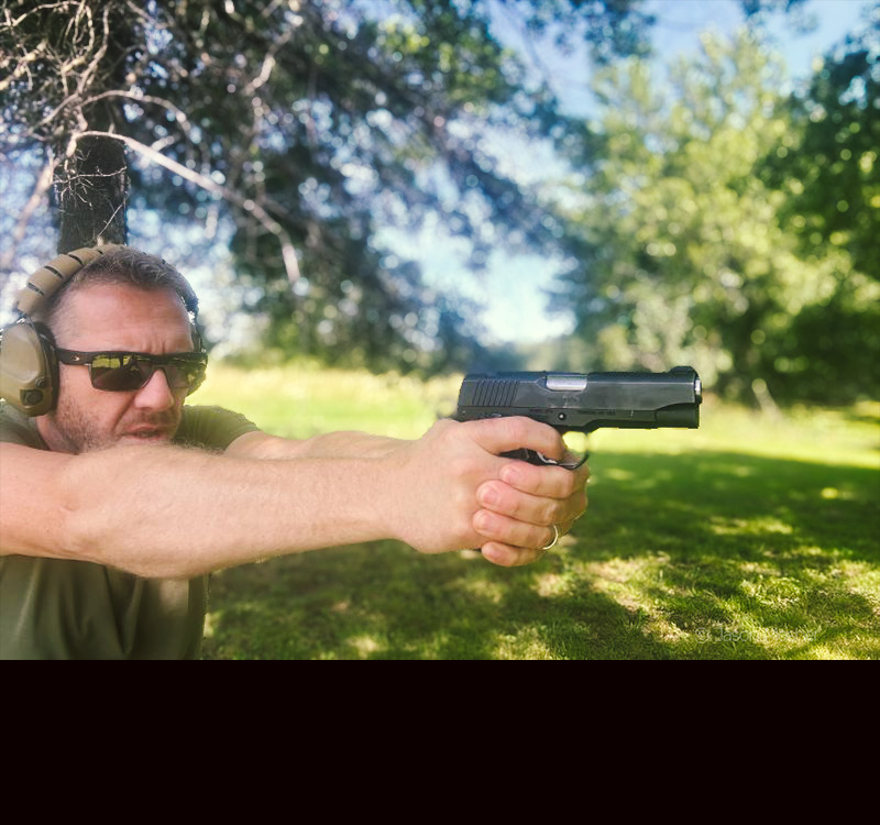 A man in a green shirt and sunglasses, shooting a 1911 pistol. 