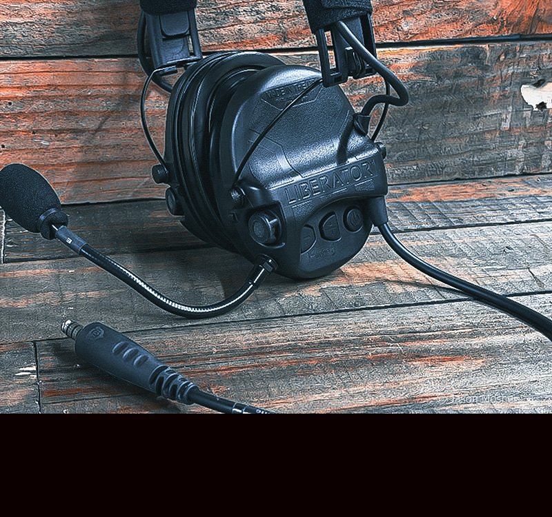 A black headset laying on a wood table with the wires for the radio coming out of the headset.