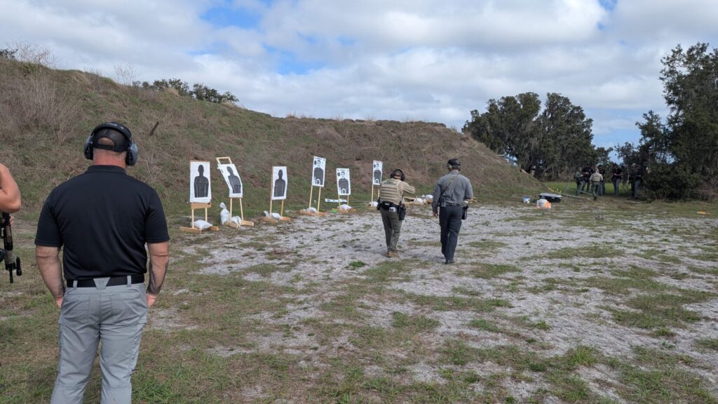 A wide shot of an instructor leading a tactical firearms class at an outdoor range, with officers observing from the left.