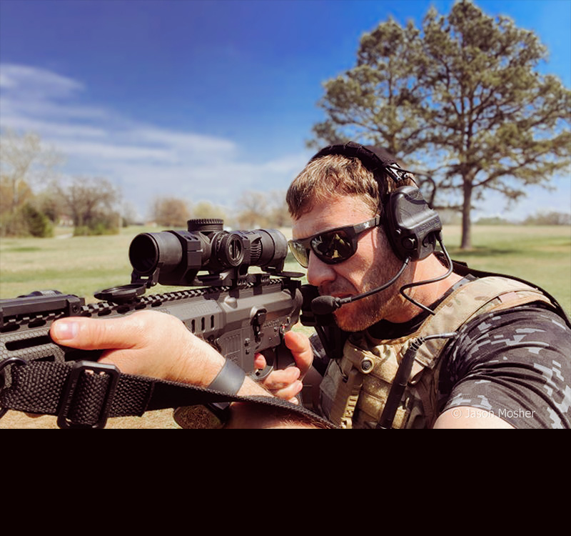 A person in a camo best and sunglasses, shooting a rifle with while wearing the Liberator IV Comms headset.