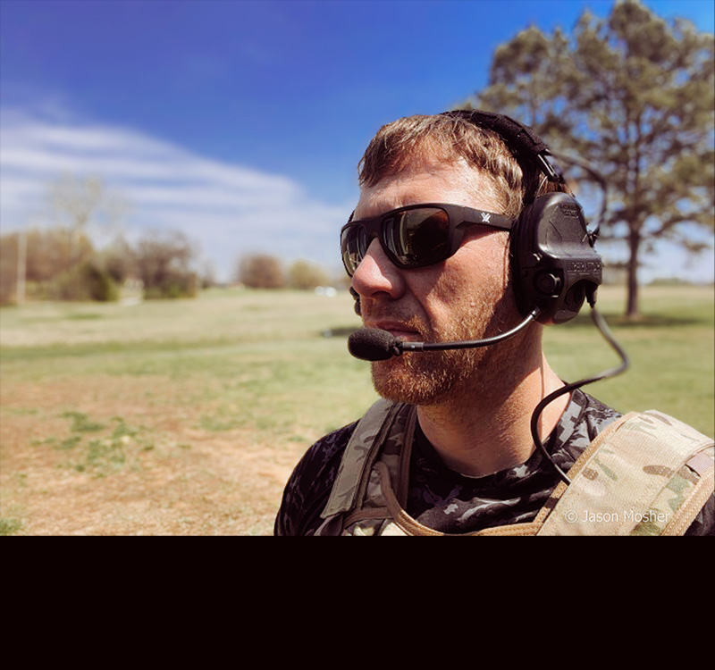 A person in a camo vest and sunglasses wearing the Liberator headset while standing in a field.