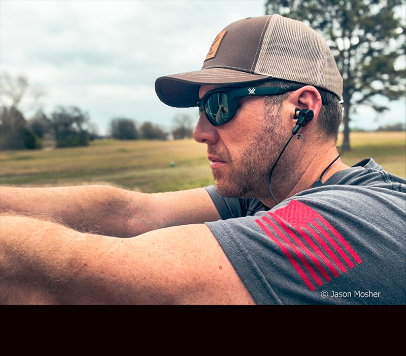 A person in a gray shirt, shooting a handgun with the PRO Impulse ear buds on.