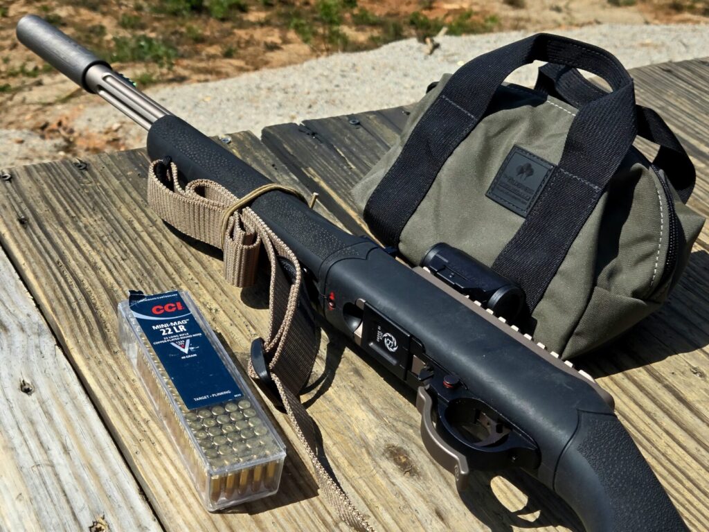 A box of ammunition with a rifle and a small bag on a wooden rifle bench