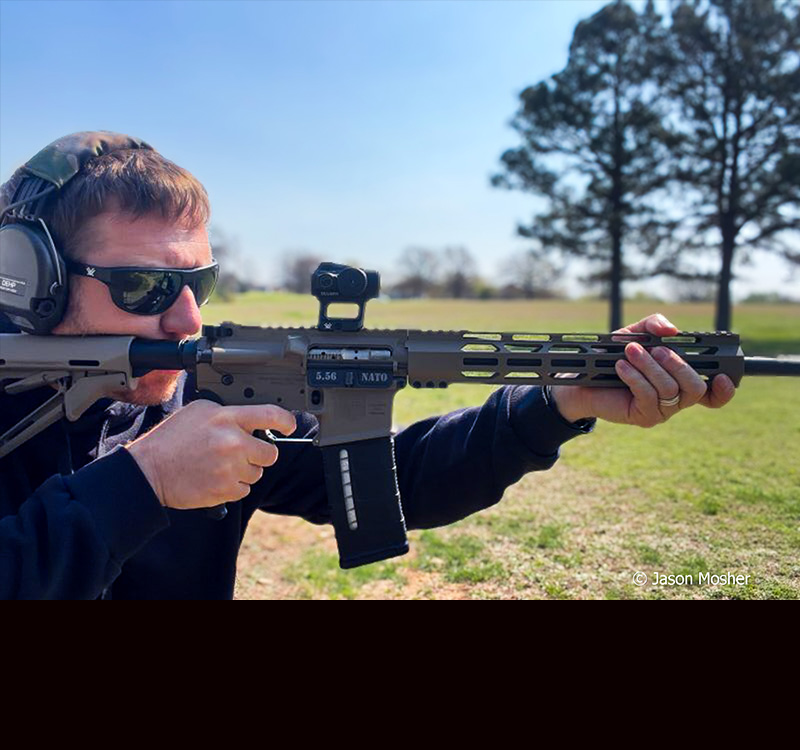 A person shooting a brown rifle with a black red dot attached to it. 