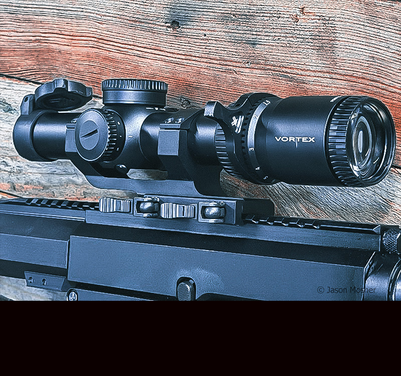 A black scope mounted on a black rifle, leaning up against a wood table.
