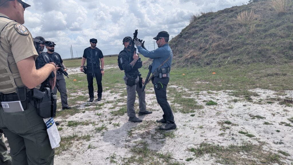 A close-up view of an instructor presenting a pistol to officers, with a series of "Warhog" brand training targets visible in the background.