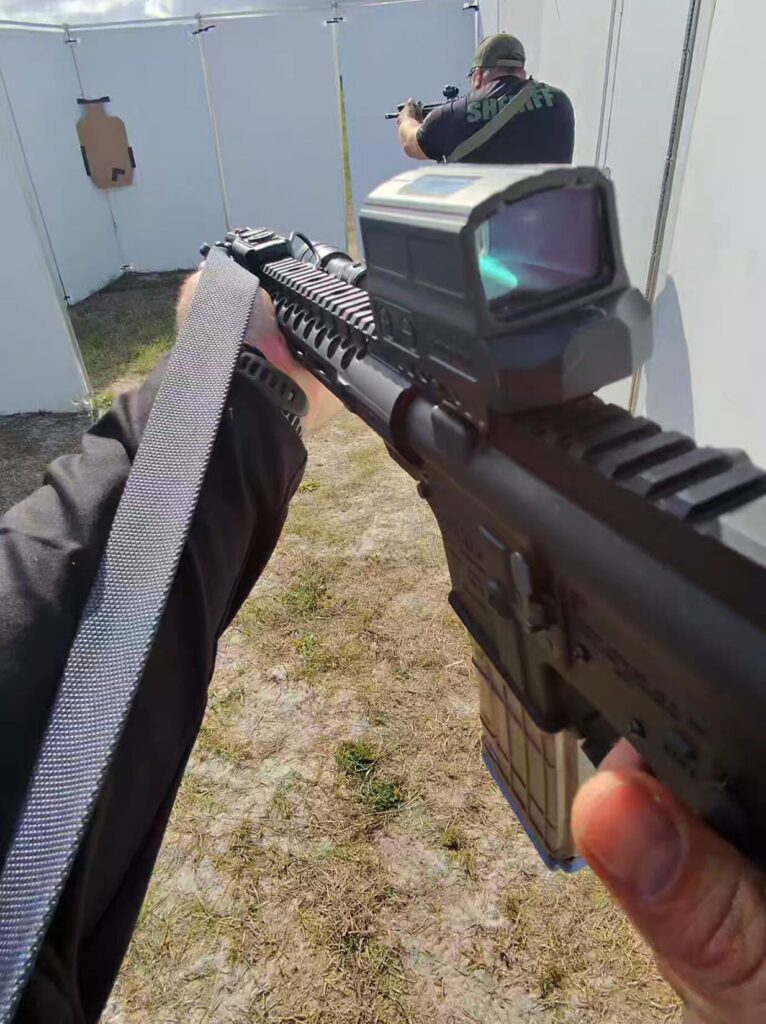 Point-of-view shot down the rail of a rifle with a Magpul MS1 sling, another shooter visible ahead