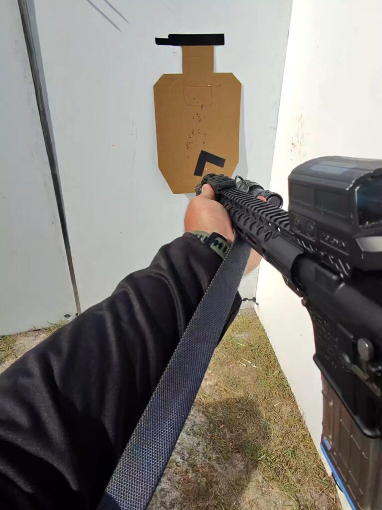 A first-person view of a shooter aiming a rifle equipped with a grey Magpul MS1 sling at a cardboard target.