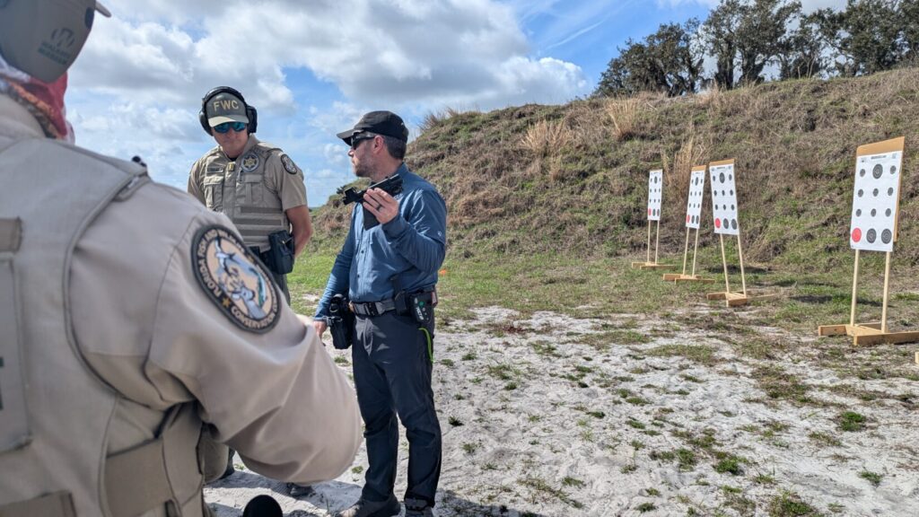 An instructor uses hand gestures to explain a shooting drill to a squad of tactical officers standing on a sandy range.