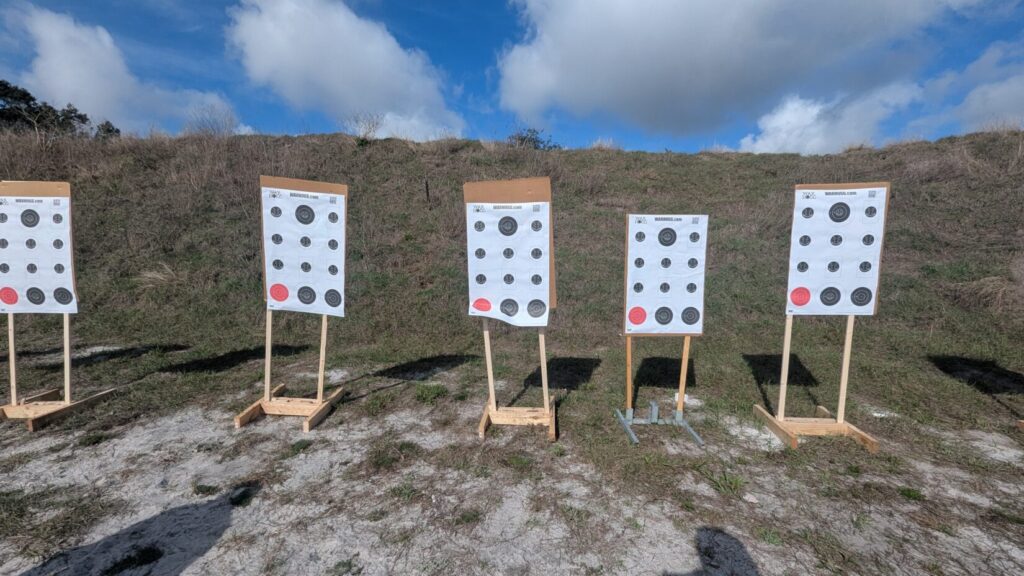 A row of multi-dot paper training targets stands on wooden frames against a grassy berm under a blue sky.
