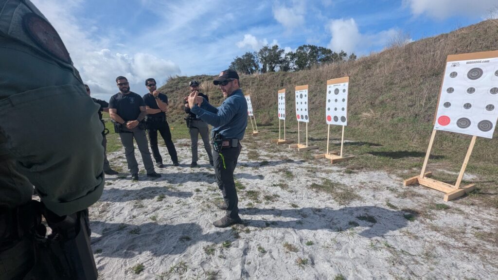 An instructor uses hand gestures to explain a shooting drill to a squad of tactical officers standing on a sandy range.