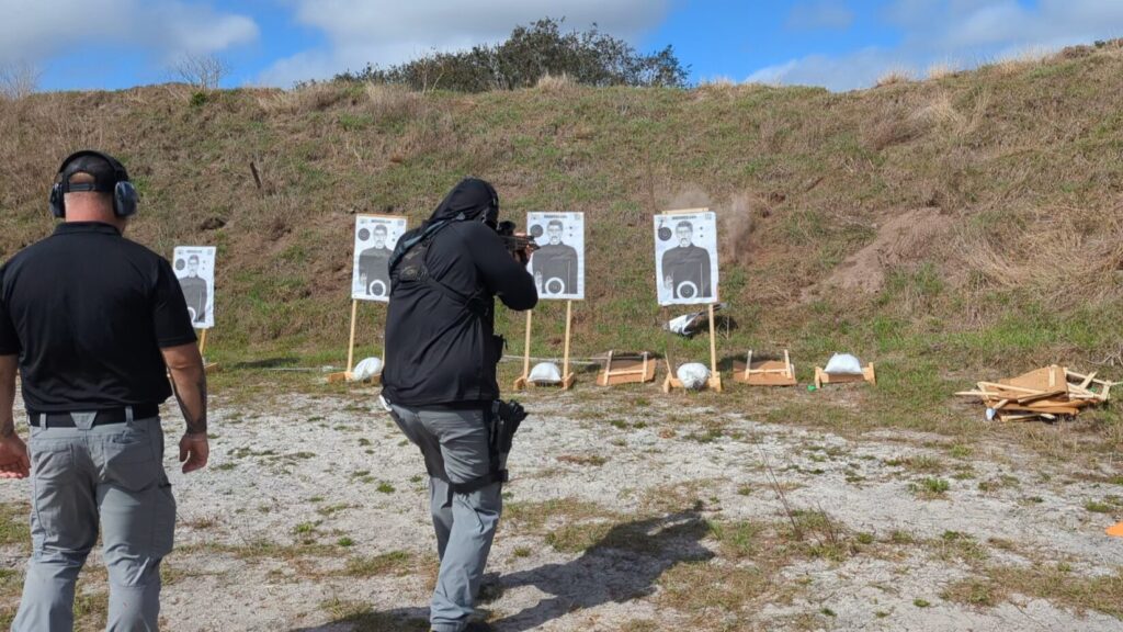 An outdoor range scene showing a shooter in a black sun hoodie engaging humanoid paper targets while an instructor observes from the side.