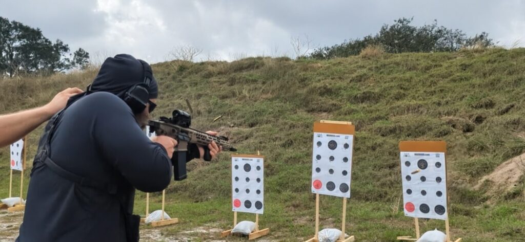 A shooter wearing the Viktos Taculus chest rig while firing a rifle at an outdoor range, with brass ejecting and several paper targets in the foreground.