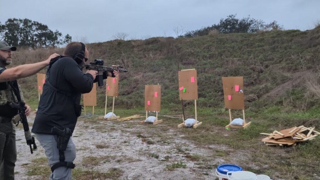 An over-the-shoulder view of a shooter aiming a suppressed MCX-SPEAR LT at cardboard targets with pink aiming points.