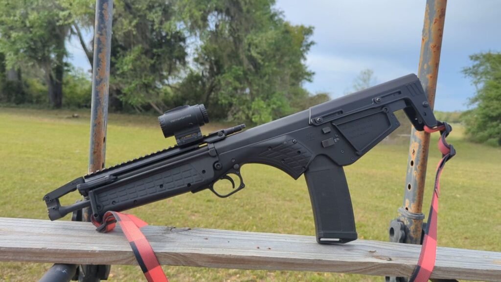 A side-view of a shooter aiming the KelTec RDB-S, showcasing the mounted prism sight and the rifle's compact bullpup layout.