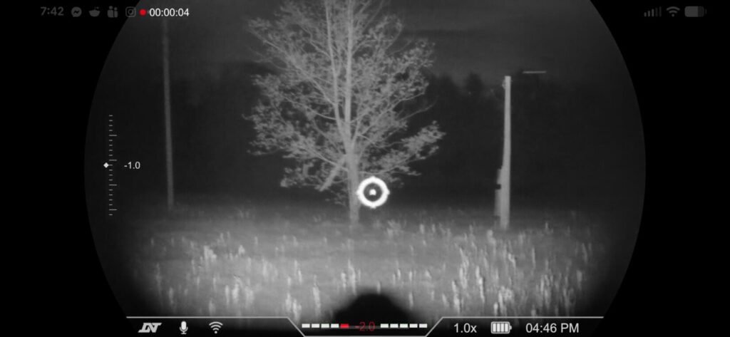 Digital night vision view of a field at night, showing a tree and power pole with a circular reticle and status icons for battery and zoom.