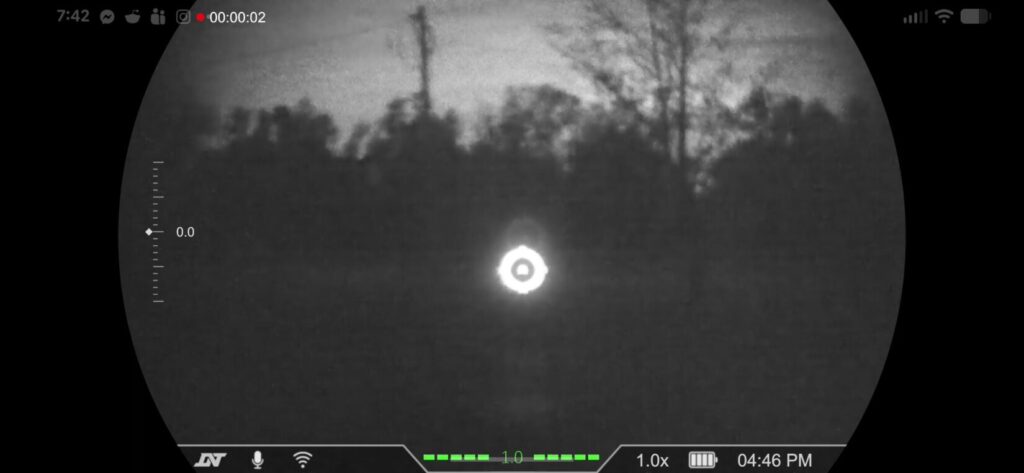 Digital night vision view of a field at night, showing a tree and power pole with a circular reticle and status icons for battery and zoom.