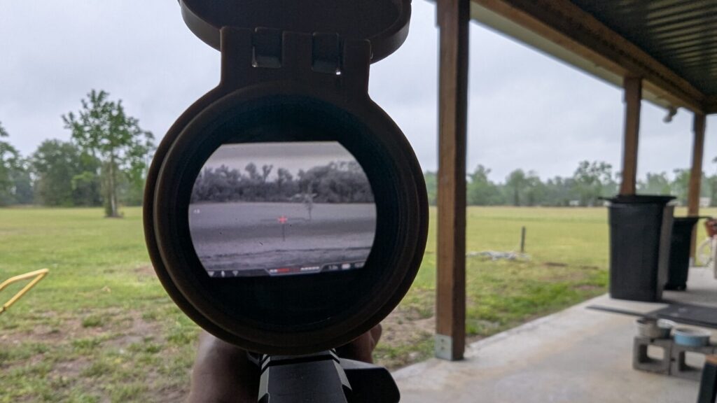 Through-the-optic view showing the digital grayscale night vision display of the NVMD-C200 with a red reticle overlay.