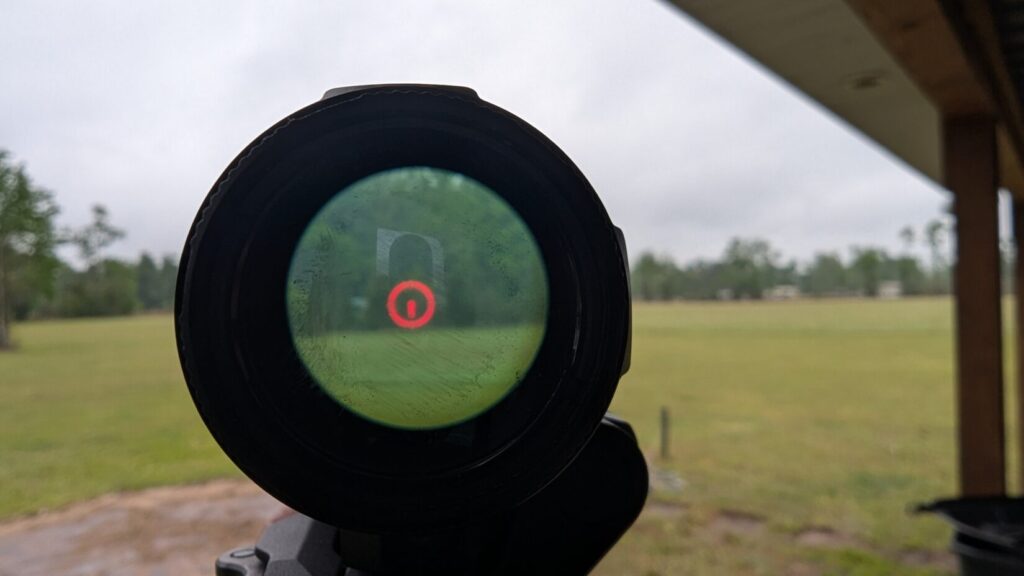 A point-of-view shot through the magnifier, showing the red circle-dot reticle focused on a target in a field.
