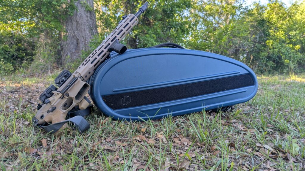 A tan rifle leaning against the closed blue Savior Pro Touring case on a grassy field.
