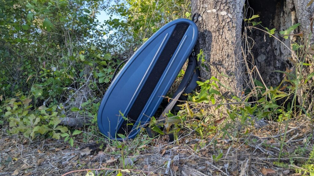 The blue Savior rifle case leaning vertically against a tree trunk in a wooded area.