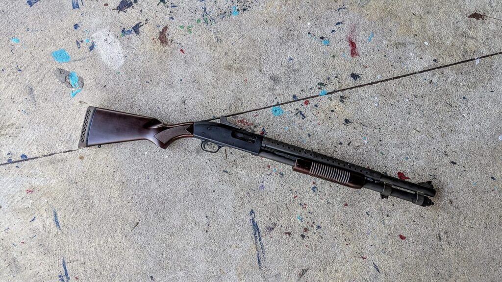  An angled overhead shot of the full shotgun lying on a concrete surface, showcasing its classic "trench gun" aesthetic with wood furniture and a heat shield.