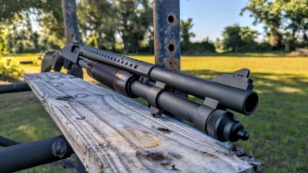 A close-up of the front half of a Mossberg M90A1 shotgun, showing the ribbed wooden corncob forend, the ventilated heat shield, and the extended magazine tube.