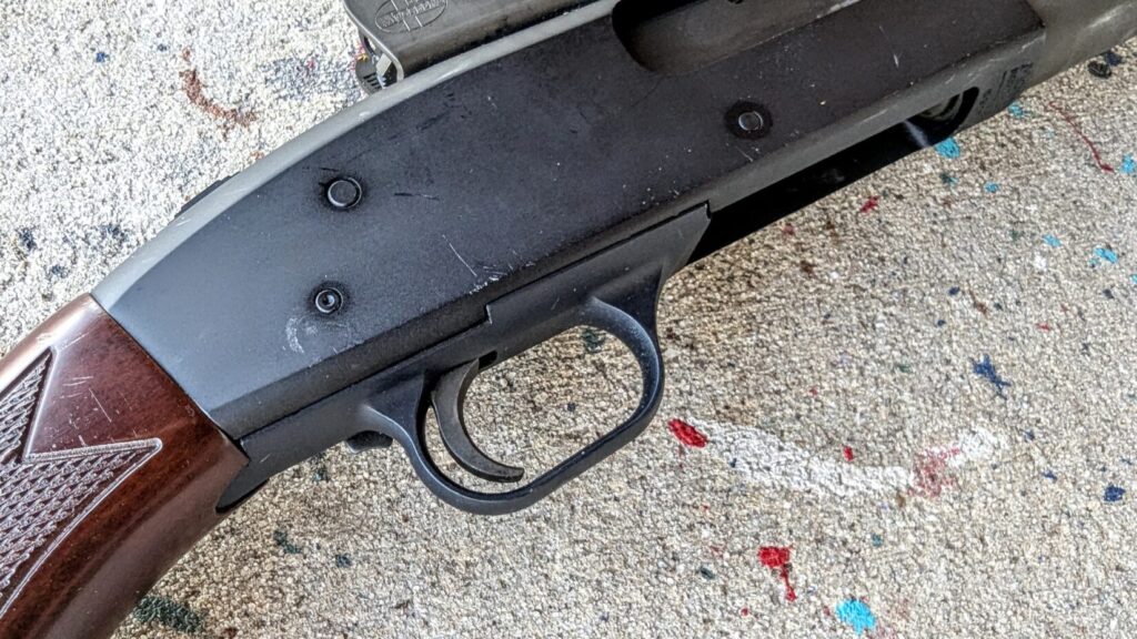 A close-up of the trigger guard and receiver area of the shotgun, showing the matte black finish and the textured grip of the wooden stock.