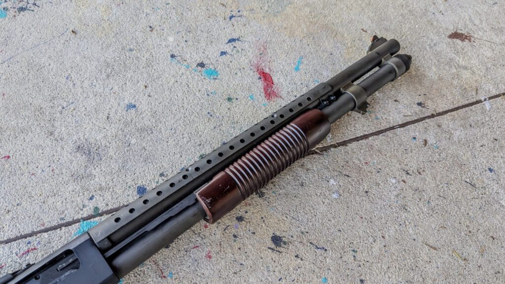 A top-down view of the Mossberg 590A1 shotgun, highlighting the heat shield over the barrel and the dark wood furniture against a gray floor.